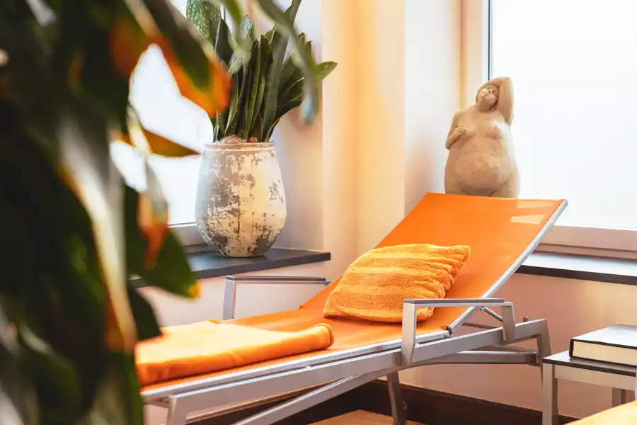 Liegefläche mit orangefarbenem Handtuch im hellen Ruheraum, umgeben von Pflanzen und warmem Licht.