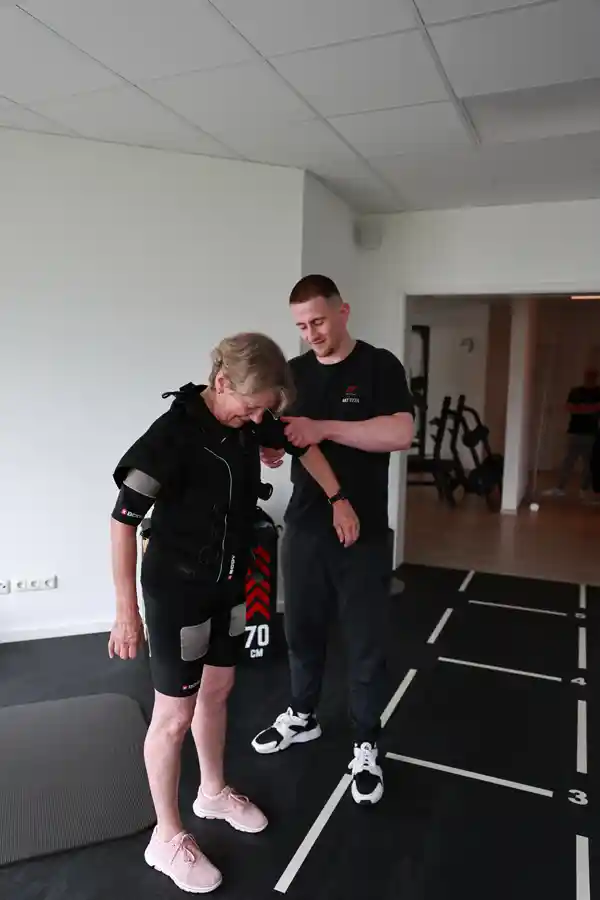 EMS-Training Vorbereitung Trainer unterstützt eine Frau beim Einstieg ins EMS-Training auf einer markierten Trainingsfläche.