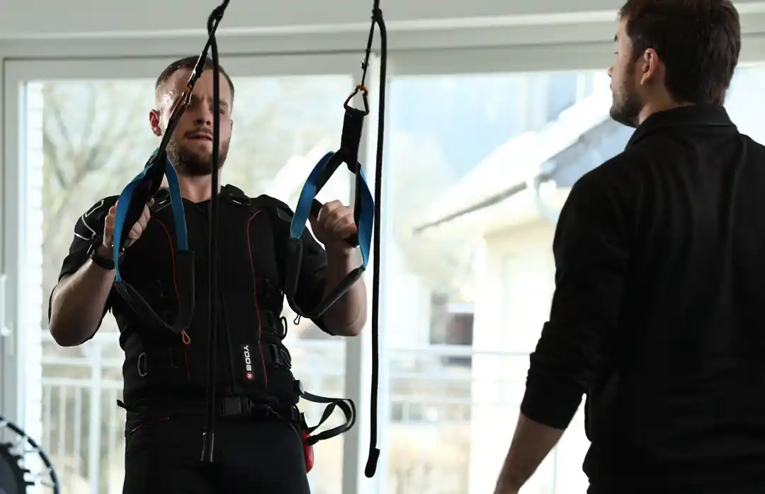 Mann beim EMS-Training mit angelegter Elektrodenweste und Gurtsystem, angeleitet von einem Trainer in moderner Trainingsumgebung.